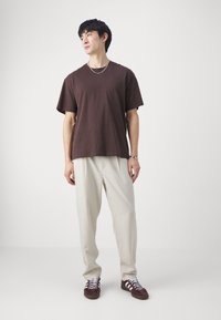 T-shirt en coton marron à manches courtes, associé à un pantalon beige clair. La tenue est complétée par des baskets bordeaux et une chaîne en argent.