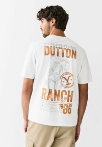 Valge lühikeste varrukatega T-särk, millel on graafiline print "Yellowstone Dutton Ranch" oranžis ja hallis värvitoonis ning hobusõitja illustratsioon.