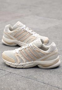 Paar beige und weiße Mesh-Laufschuhe mit weißen Schnürsenkeln auf grauer gefliester Bodenfläche.