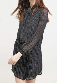 Blusa nera trasparente con pois bianchi, maniche lunghe e scollo con fiocco. Presenta una vestibilità comoda e un orlo leggermente arrotondato.