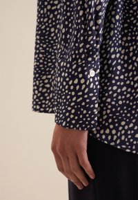 Blusa blu navale con motivo a pois bianchi, maniche lunghe, dettaglio con bottone sui polsini, tessuto morbido e vestibilità comoda. Primo piano della manica e della mano.