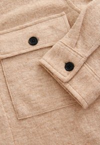 Beige villakangas, jossa on pehmeä tekstuuri, ja jossa on päällysläpä tasku sekä hihassa kaksi mustaa nappia. Yksinkertainen, klassinen muotoilu.