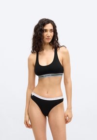 Woman with curly dark hair wearing black sports bra and matching briefs with white branded waistbands, standing against plain light background.