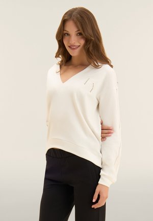 Giovane donna con capelli mossi che indossa un maglione bianco con scollo a V e pantaloni neri, sorridente con un braccio dietro la schiena.