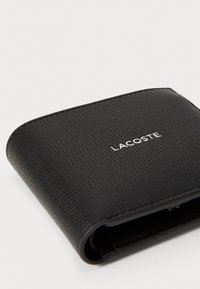 Svart lær lommebok med teksturert overflate, med rundede hjørner og en hvit trykt "LACOSTE"-logo på forsiden.