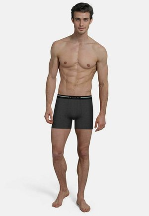 Junger Mann mit kurzen dunklen Haaren steht barfuß in dunkelgrauen Boxershorts mit "bugatti"-Bund vor weißem Hintergrund.