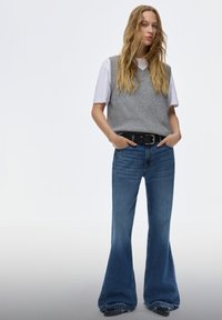 Mladá žena s dlhými blond vlasmi, ktorá má na sebe šedú pletenú vestu cez biele tričko a modré nohavice s vysokým pásom a rozšírenými nohavicami, pričom nosí čierny opasok.