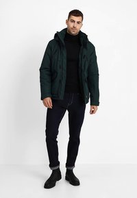 Tummanvihreä toppatakki hupulla, jossa on sileä pinta, mustia yksityiskohtia ja etuvetoketjut. Päällä musta pooloneule ja tummat farkut.