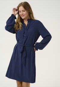 Robe bleu marine à manches longues, avec plastron à boutons froncés et ceinture à nouer à la taille. Tissu doux avec une texture lisse. Design longueur sol.