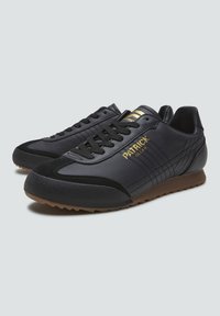 Par de zapatillas negras de corte bajo con suelas marrones, cordones negros y la marca dorada "Patrick Milan" en el costado.