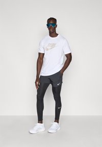 Fehér Nike póló, rajta bézs "RUN" felirattal, fekete rövidnadrág, fekete leggings és fehér sportcipő. Kék napszemüveg teljesíti az atletikus megjelenést.