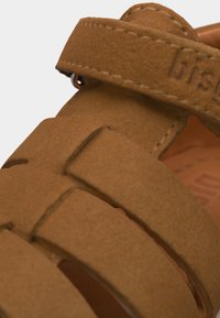 Κοντινή λήψη των καφέ σουέντ λουριών των σανδαλιών με ορατή ραφή και μερικά ανάγλυφα γράμματα με το όνομα της μάρκας στο ρυθμιζόμενο λουράκι στον αστράγαλο.