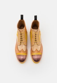 Botas de cuero bicolor con una mezcla de tonos beige, amarillo y naranja. Presentan detalles de brogue y una puntera de cobre redondeada y desgastada.