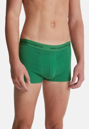 Mann in grünen Boxershorts mit einem Bund von "United Colors of Benetton", der vor einem schlichten weißen Hintergrund steht.