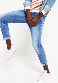 Jeans de mezclilla azul claro con un corte ajustado, puños enrollados y una textura desgastada, combinados con zapatillas blancas. Camisa de rayas visible arriba.