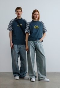 Δύο μοντέλα στέκονται μαζί. Και οι δύο φορούν φαρδιά, σκούρα μπλε μπλουζάκια με μανίκια σύντομων σχεδίων και φαρδιά, ανοιχτό μπλε τζιν παντελόνια.