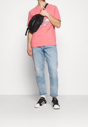 Schwarze Umhängetasche mit verstellbarem Tragegurt, mit weißem Logo, getragen über einem rosa Hemd und kombiniert mit hellblauen Jeans und schwarzen Sneakers.