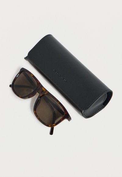 Ορθογώνια γυαλιά ηλίου tortoiseshell με σκούρα φακούς δίπλα σε μαύρη δερμάτινη θήκη Dolce & Gabbana σε λευκό φόντο.