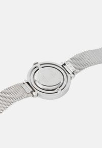 Srebrny zegarek z okrągłą kopertą i siateczkowym paskiem; charakteryzuje się gładkim, polerowanym wykończeniem oraz widocznym logo marki na tylnej stronie.