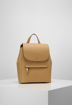 Sac à dos en cuir fauve avec poignée supérieure, fermeture à rabat et poche zippée à l'avant, posé sur un piédestal blanc contre un fond gris uni.
