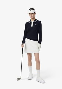 Polo à manches longues en maille bleu marine avec col blanc, jupe blanche, visière blanche et club de golf. Des baskets blanches et un gant complètent le look sportif.