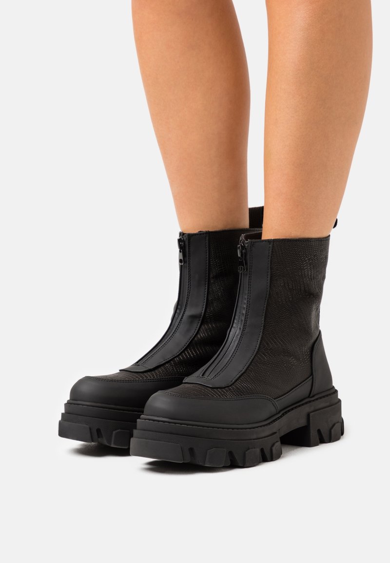Bottes à plateforme épaisses noires mi-mollet avec fermetures éclair avant, portées par une personne aux jambes nues sur fond blanc.