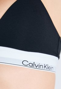 Primo piano di una persona che indossa un reggiseno sportivo nero di Calvin Klein con un elastico bianco e testo del logo nero.