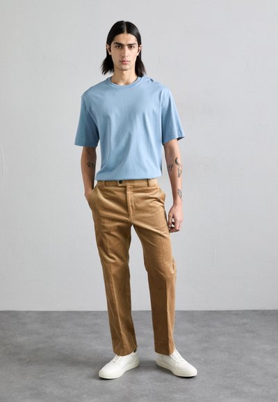 Camiseta de algodón azul claro con mangas cortas, combinada con pantalones de pana marrón y zapatillas blancas. Diseño limpio, textura suave, ajuste relajado.