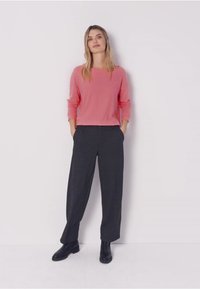 Nainen seisoo valkoista seinää vasten pukeutuneena pitkähihainen vaaleanpunainen paita, löysät tummanharmaat housut ja mustat nilkkurit, kädet taskuissa.