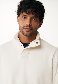 Sudadera color crema con un cuello alto, presenta un cierre de botones a presión, un logotipo sutil y una tela suave y texturizada.