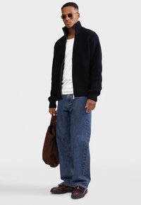 Dunkelblauer, gerippter Cardigan mit hohem Kragen und Reißverschluss. Weißes T-Shirt darunter. Blaue, locker sitzende Jeans. Braune Tasche und bordeauxrote Loafers.