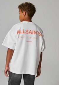 Bílý oversized tričko s potiskem na zádech v oranžovém textu, včetně nápisů "ALLSAINTS" a "LONDON", s kulatým výstřihem a krátkými rukávy.