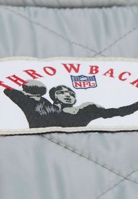 Graumelierter Stoff mit einem Patch, auf dem in roter Schrift "THROWBACK", das NFL-Logo und eine schwarz-weiße Grafik eines Spielers, der einen Football wirft, zu sehen sind.