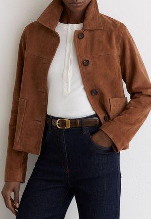 Person wearing a brown suede button-up jacket over a white ribbed henley shirt, dark blue jeans, and a brown belt with a gold buckle.