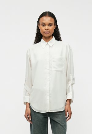 Chemise blanche à boutons, en tissu léger, avec une poche, coupe décontractée et manches retroussées, associée à un jean gris.