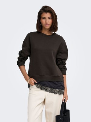 Ung kvinde iført en mørk langærmet sweater, hvide bukser, holder en sort taske, med en sort blonderet top, der titter frem under.