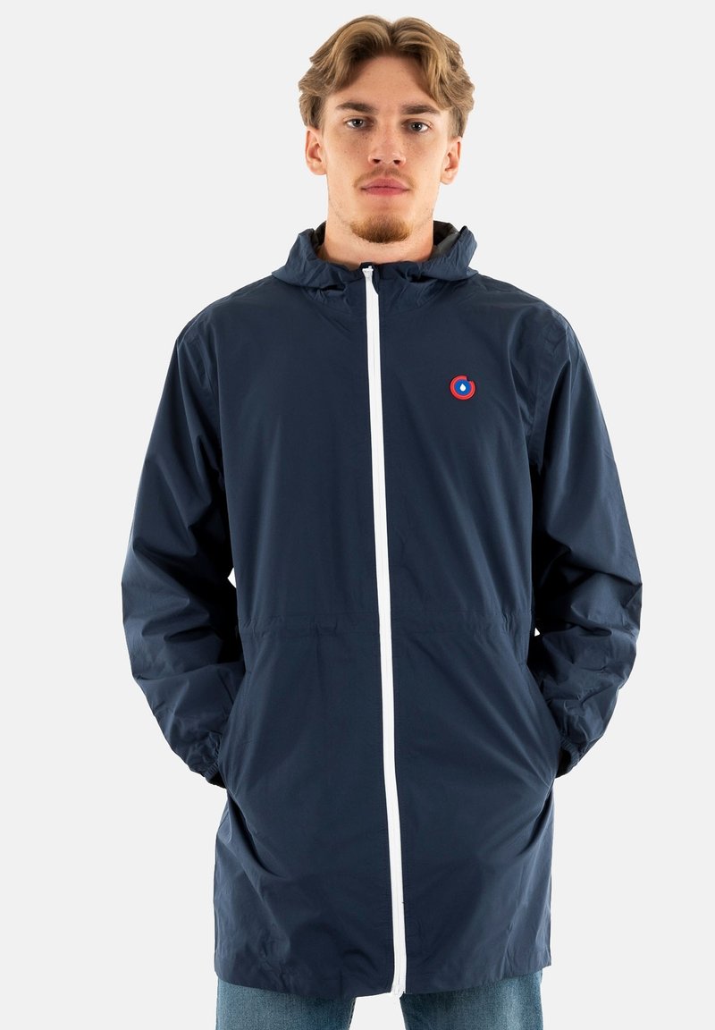 Veste imperméable légère marine avec capuche, fermeture éclair blanche et un logo rouge circulaire sur la poitrine. Elle possède des poignets élastiques et une coupe décontractée.