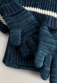 Strikkede handsker i mørk teal med ribbede manchetter, der har et tekstureret mønster og lysere blå striber, placeret ovenpå en matchende sweater.
