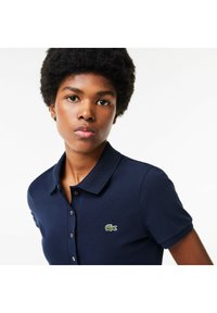 Mörkblå pikétröja med krage och korta ärmar, med en grön Lacoste-logotyp på vänster bröst. Texturerad tyg med knäppning.