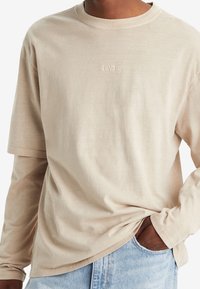 Beżowa koszula Levi's z długim rękawem nałożona na beżowy T-shirt z krótkim rękawem, noszona z jasnoniebieskimi dżinsami, ręka w kieszeni.