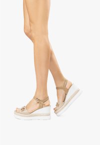 Sandali platform beige con cinturino alla caviglia, una fibbia decorativa e un dettaglio in corda intrecciata su una suola a zeppa bianca. Texture liscia e design aperto sulla punta.
