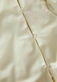 Tessuto beige con una texture liscia e leggermente lucida, presenta uno spacco e piccole cuciture lungo la cucitura per un tocco di dettaglio.