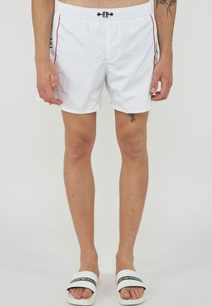 Man draagt witte zwembroek met rode bies en zwart-witte Emporio Armani slippers, met zicht op been- en tatoeëer details.