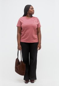 Donna con i capelli intrecciati guarda verso destra, indossa una maglietta rosa con piccoli dettagli a forma di cuore, pantaloni neri e tiene in mano una borsa shopper marrone.