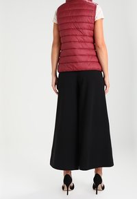 Femme debout, tournée vers l'arrière, portant un gilet matelassé rouge sans manches, un haut blanc à manches courtes, un pantalon large noir et des escarpins noirs.