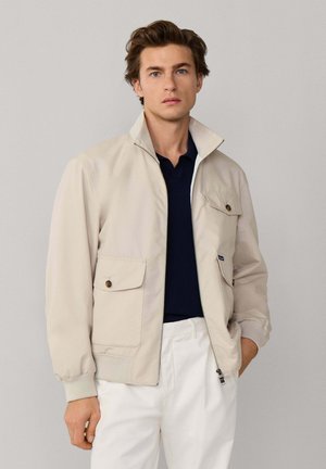 Joven con chaqueta beige con cremallera sobre una camisa polo azul marino oscuro y pantalones blancos, de pie frente a un fondo liso de color gris claro.