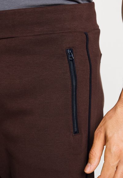 Calças de treino castanhas com uma cintura justa, bolso lateral com fecho e detalhe em fecho éclair azul escuro em contraste. Tecido macio e texturizado.