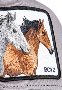 Pilka kepurė su stačiakampiu užtrauktuku, kurioje yra siuvinėti pilko ir rudo arklio galvos, prieš baltą foną, su užrašu "BOYZ".