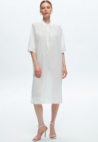 Robe en coton blanc avec un col et un décolleté à nouer, manches courtes featuring des détails en œillet, et une silhouette ample et décontractée.
