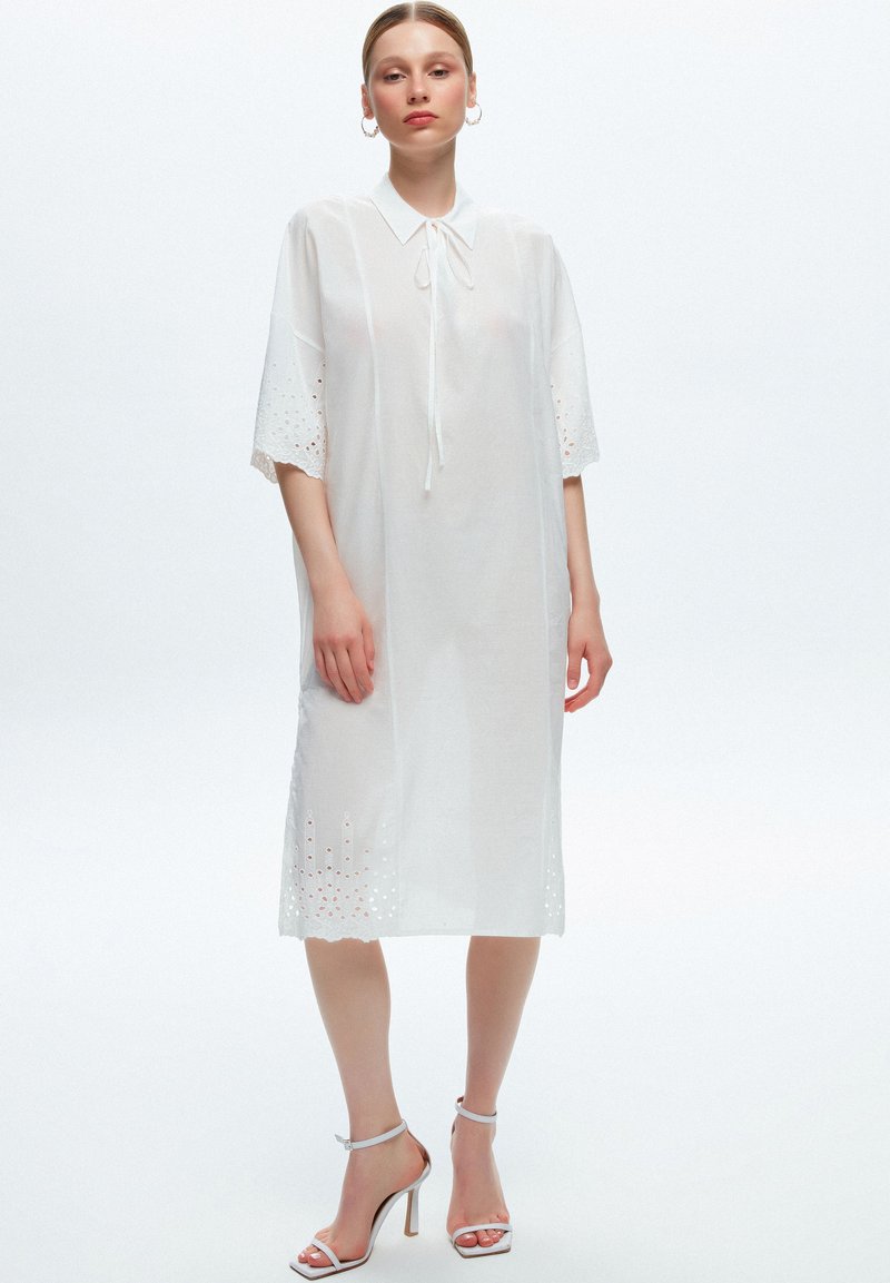 Robe en coton blanc avec un col et un décolleté à nouer, manches courtes featuring des détails en œillet, et une silhouette ample et décontractée.